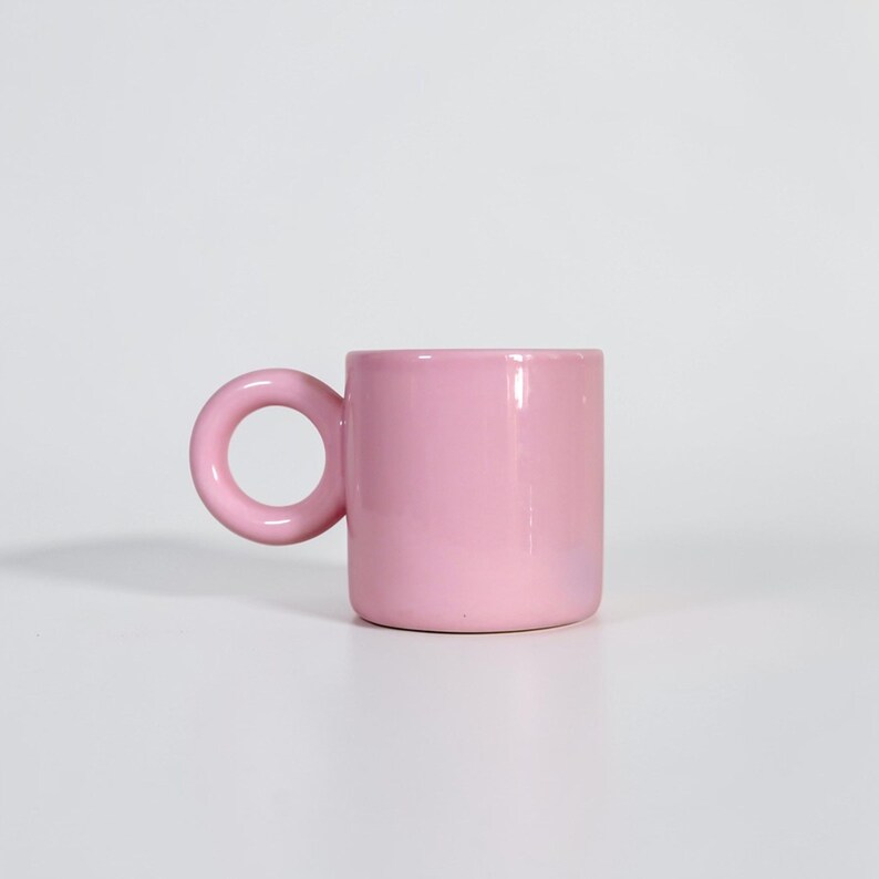 May include: A solid pink ceramic mug with a rounded handle. The mug has a cylindrical body and a smooth, glossy finish. The handle is a large, circular loop. The mug is set against a white background.