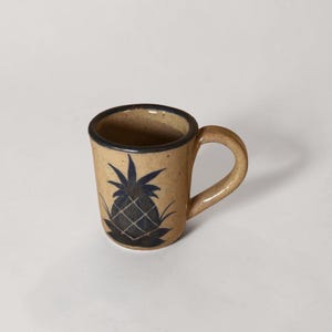May include: A small, tan ceramic mug with a dark blue pineapple design. The mug has a rounded handle and a dark blue rim. The pineapple design is centered on the front of the mug.