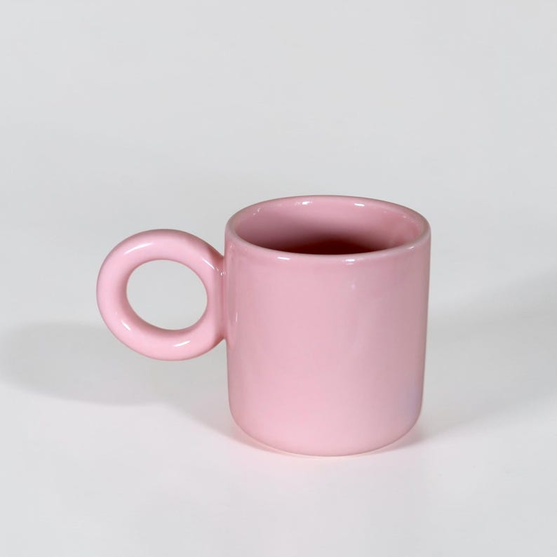May include: A solid pink ceramic mug with a rounded handle. The mug has a cylindrical shape and a smooth, glossy finish. The handle is a circular loop, and the interior of the mug is also pink.