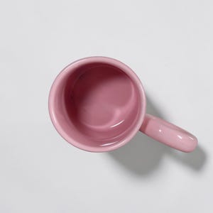 May include: A top-down view of a solid pink ceramic mug with a matching handle. The mug has a smooth, glossy finish and a simple, rounded design. The interior of the mug is also pink.