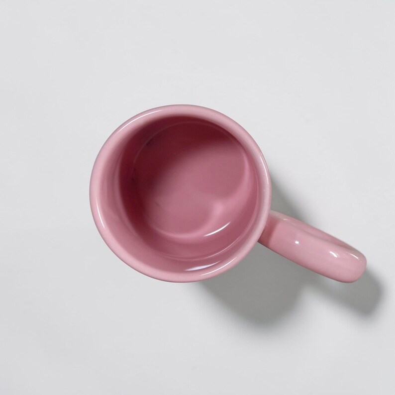 May include: A top-down view of a solid pink ceramic mug with a matching handle. The mug has a smooth, glossy finish and a simple, rounded design. The interior of the mug is also pink.