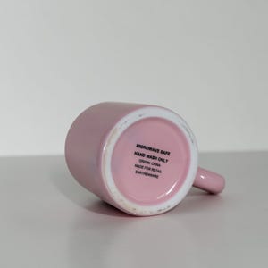 May include: A light pink ceramic mug with a handle, lying on its side. The bottom of the mug is visible, displaying text that reads "MICROWAVE SAFE", "HAND WASH ONLY", "ORIGIN: CHINA", "MADE FOR RETAIL", and "EARTHENWARE".