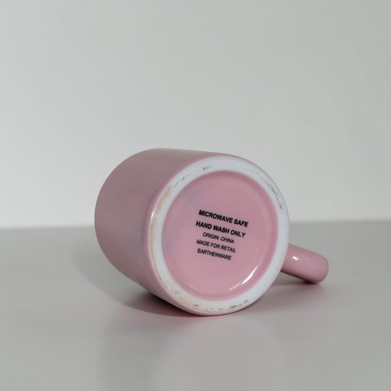 May include: A light pink ceramic mug with a handle, lying on its side. The bottom of the mug is visible, displaying text that reads "MICROWAVE SAFE", "HAND WASH ONLY", "ORIGIN: CHINA", "MADE FOR RETAIL", and "EARTHENWARE".