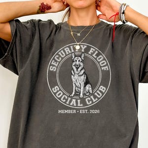 May include: Dark grey t-shirt featuring a circular graphic with a German Shepherd dog illustration. The text around the dog reads "SECURITY FLOOF SOCIAL CLUB" with "MEMBER • EST. 2026" below. The shirt has a vintage, washed look.