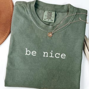 May include: An olive green t-shirt with the words "be nice" in white block letters. A gold necklace with a small pendant rests on the shirt. The shirt is folded and next to a brown hat.