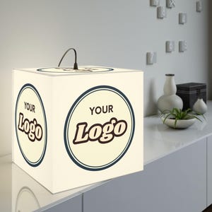 Może przedstawiać: Duża, podświetlana kostka z napisem "YOUR Logo" w okrągłym wzorze na bokach. Kostka ma wymiary 30 x 30 x 30 cm i uchwyt na górze. Kostka stoi na białej powierzchni.