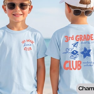 May include: A light blue t-shirt featuring a small graphic on the front with the text 3rd GRADE Jamie CLUB. The back displays a larger graphic with the text 3rd GRADE Level Up!! CLUB memberships unlocked, alongside illustrations of a paper airplane, books, and a star.