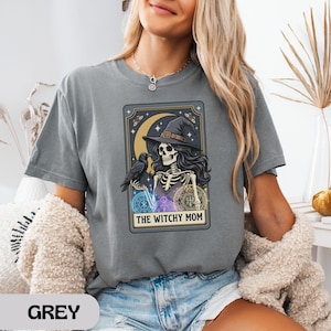 Woman wearing grey t-shirt with witchy skeleton graphic and text.