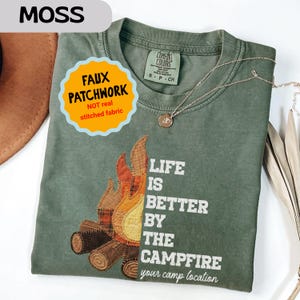 May include: Moss green t-shirt with a faux patchwork campfire design. The shirt features the text "LIFE IS BETTER BY THE CAMPFIRE" and "your camp location". A gold necklace is draped across the shirt.