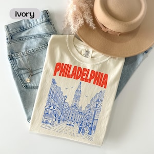 May include: An ivory t-shirt featuring the word PHILADELPHIA in bold red letters above a blue line art illustration of a city street scene with a pretzel cart, a tall clock tower, and people walking, paired with light blue denim jeans and a tan hat.