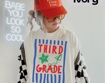 May include: An ivory t-shirt featuring a graphic with blue vertical stripes, red text reading THIRD GRADE, a green star, and a green paper airplane. The outfit includes an orange trucker hat with the text MAD LOVE FOR YOU in white letters.