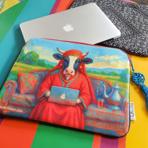 May include: A colorful laptop sleeve featuring a cow dressed in red robes, holding a laptop with an apple logo. The sleeve has a blue zipper and a blue tassel. A silver laptop rests on top of the sleeve.