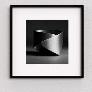 May include: A black and white framed print featuring a metallic cuff bracelet with a brushed finish. The bracelet has a distinct twisted, overlapping design that creates a continuous loop, captured in a studio setting with dramatic lighting.