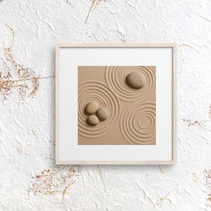May include: A framed print featuring a zen garden design. The image shows a tan sand surface with concentric circles raked into it. Three small stones are clustered on the left, and one larger stone is on the right. The frame is light-colored.