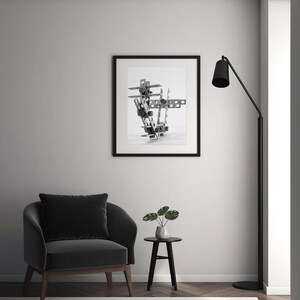 May include: A black and white framed print of a metal sculpture. The sculpture is abstract and geometric, with various shapes and angles. The print hangs above a black armchair with a matching pillow. A small side table with a plant and a black floor lamp are also in the room.