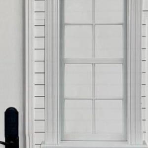 Op de afbeelding: Een gedeelte van een witte buitenmuur met een raam en een deur. Het raam heeft een rechthoekig frame en een rasterpatroon. De deur heeft een zwarte handgreep. De muur heeft horizontale gevelbekleding.