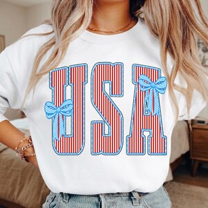 May include: White sweatshirt with the word "USA" in red and white striped letters with blue outlines. The letters have a pattern of small white stars and are decorated with blue bows with white polka dots. A patriotic theme.