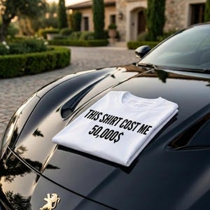May include: A white t-shirt with black text printed on the front that reads, "THIS SHIRT COST ME 50,000$". The garment is folded and placed on the hood of a black sports car, which features a gold emblem near the front edge of the vehicle.