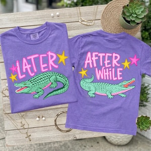 May include: Two purple t-shirts with a green alligator graphic. One shirt says "LATER" with pink and yellow accents, and the other says "AFTER WHILE". The alligator has a red mouth and is surrounded by stars.