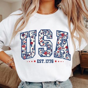 May include: White sweatshirt with the word "USA" in a blue outline, filled with a floral pattern of red flowers and blue leaves. Below the word "USA" is the text "EST. 1776" with red stars on each side. A patriotic themed top.
