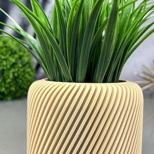 May include: A cylindrical beige planter with a ridged, diagonal design, filled with vibrant green grass-like plants. The planter is made of a light-colored material and sits on a gray surface. The overall theme is home decor.