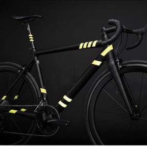 May include: A sleek black road bicycle with yellow accent stripes on the frame and seat post. The bicycle has black wheels, tires, and handlebars, set against a dark background. The bike is designed for speed and performance.