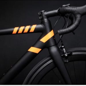 May include: A sleek black road bike with orange accent stripes on the frame. The handlebars are wrapped in black tape, and the tires are black. The bike is set against a dark background.