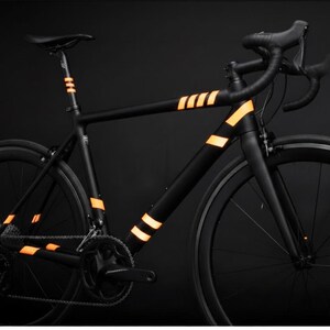 May include: A sleek black road bike with orange reflective stripes on the frame and seat post. The bike has black wheels and handlebars, set against a dark background. The design emphasizes a modern, minimalist aesthetic.