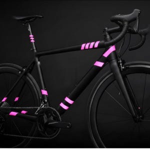 May include: A sleek black road bike with vibrant pink accents. The bicycle features a matte black frame with pink stripes on the frame and seat post. The wheels and components are black, creating a striking contrast against the pink details. A modern and stylish design.