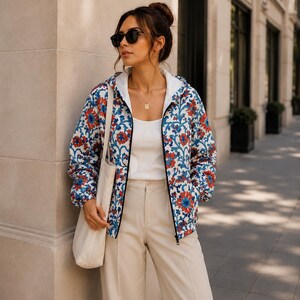 May include: A woman wearing a white tank top, beige pants, and a floral print jacket with red and blue flowers. She is also wearing sunglasses and carrying a beige tote bag. The jacket has a black zipper.