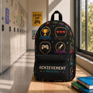 May include: A black backpack featuring a repeating pixel art pattern of video game icons including trophies, hearts, swords, and controllers. The front pocket displays the text "ACHIEVEMENT UNLOCKED" in a pixelated font style with a black background.