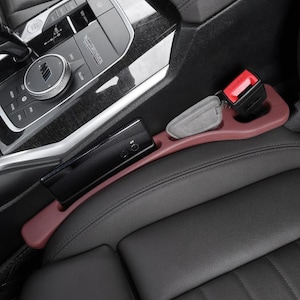 Puede incluir: Un organizador de espacio para asiento de coche en color granate, situado entre un asiento de cuero negro y la consola central. El accesorio cuenta con una ranura alargada para un tel&eacute;fono m&oacute;vil y un hueco circular para una llave gris.