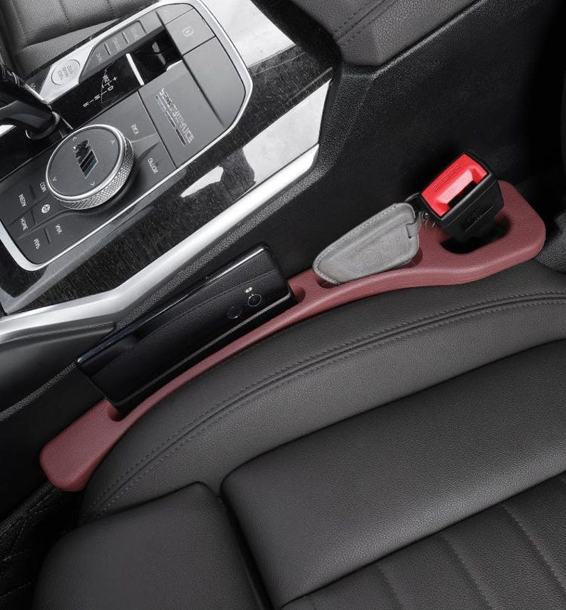 Puede incluir: Un organizador de espacio para asiento de coche en color granate, situado entre un asiento de cuero negro y la consola central. El accesorio cuenta con una ranura alargada para un tel&eacute;fono m&oacute;vil y un hueco circular para una llave gris.