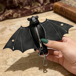 May include: Black bat-shaped coin purse with a zipper closure and silver-tone hardware. The wings are detailed with white stitching and gold rivets. A silver clasp is attached for easy carrying. The purse is held by a hand with green nail polish.