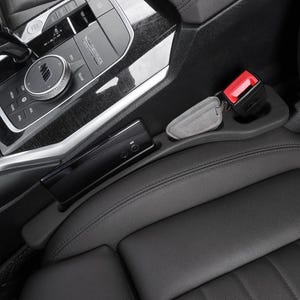Puede incluir: Un organizador de huecos para asiento de coche negro hecho de material liso, colocado entre la consola central y el asiento de cuero. Cuenta con una ranura para smartphone y un hueco para el cintur&oacute;n de seguridad, con un llavero gris dentro.