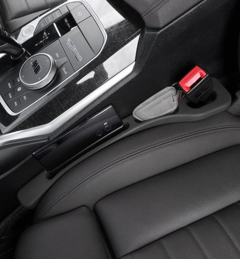 Puede incluir: Un organizador de huecos para asiento de coche negro hecho de material liso, colocado entre la consola central y el asiento de cuero. Cuenta con una ranura para smartphone y un hueco para el cintur&oacute;n de seguridad, con un llavero gris dentro.
