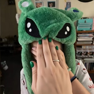 May include: A plush green hat featuring an alien character design with large black eyes, white highlights, and small ears. The hat is made of soft, fuzzy material and includes ear flaps that extend downward on both sides of the head.