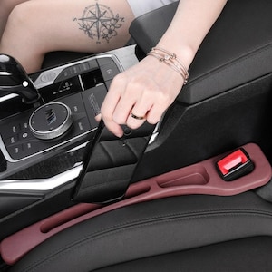Puede incluir: Un organizador de espacio para asiento de coche en color granate, hecho de un material liso y flexible. El organizador cuenta con una ranura larga y estrecha para guardar objetos y un hueco dise&ntilde;ado para encajar alrededor de un cintur&oacute;n.