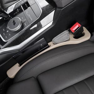 Puede incluir: Un organizador de huecos para asientos de coche en color beige, situado entre un asiento de cuero negro y la consola central. El organizador cuenta con una ranura larga que sostiene un smartphone negro y un compartimento con una llave gris.