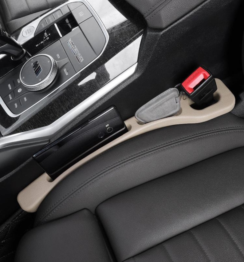 Puede incluir: Un organizador de huecos para asientos de coche en color beige, situado entre un asiento de cuero negro y la consola central. El organizador cuenta con una ranura larga que sostiene un smartphone negro y un compartimento con una llave gris.