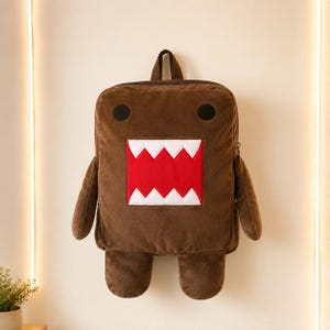 May include: A brown plush backpack shaped like a character with a rectangular body, two small arms, and two legs. The face features two black circular eyes and a large, wide-open mouth with a bright red background and sharp white teeth.