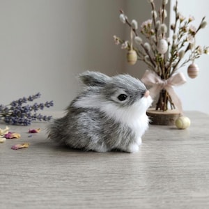 May include: A small, realistic figurine of a fluffy bunny with grey and white fur. The bunny has dark, round eyes, a small pink nose, and pointed ears. It is positioned in a seated posture, showcasing soft, textured fur and a white chest area.
