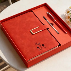 Könnte beinhalten: Ein rotes Leder-Notizbuch- und Stift-Set in einer passenden Box. Das Notizbuch hat einen goldfarbenen Verschluss und ist mit den Initialen "AR" und den Namen "Angela Rorges" geprägt. Ein silberner und roter Stift und ein schwarzer Stift sind enthalten.