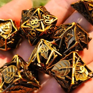 May include: A collection of gold and black metal dice featuring leaf and number detailing. The dice are held in a hand, highlighting their intricate design. The set includes various shapes, ideal for tabletop games.
