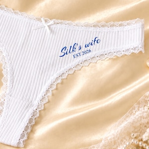 May include: White ribbed underwear featuring a delicate lace trim along the edges and a small centre bow. The fabric is printed with the blue text Silk's wife and EST.2026. The design includes a high-cut leg opening and a soft textured finish.