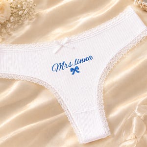 May include: A white ribbed thong featuring delicate lace trim along the edges and a small white bow at the centre waist. The front displays the blue text Mrs.linna positioned above a small blue bow graphic, creating a clean and detailed appearance.