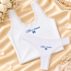 Könnte beinhalten: Ein weißes Set aus einem gerippten Tanktop und einem Tanga mit zarter Spitzenborte an den Rändern. Beide Teile sind mit dem blauen Schriftzug Mrs.linna und einer kleinen blauen Schleifengrafik in der Mitte bedruckt.