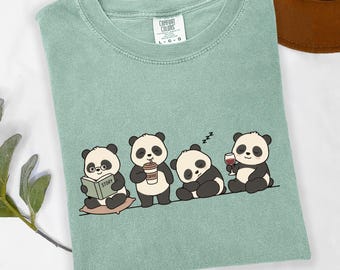 May include: A sage green crewneck t-shirt featuring a graphic of four pandas. One panda reads a book labelled STORY, one drinks coffee, one sleeps with ZZZ symbols, and one holds a glass of wine. The shirt has a Comfort Colors brand neck label.