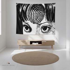 Op de afbeelding: Zwart-wit wandtapijt met een gedetailleerde illustratie van ogen en een spiraalvormig ontwerp. Het kunstwerk hangt aan een witte muur boven een houten kast en een rond, beige tapijt.