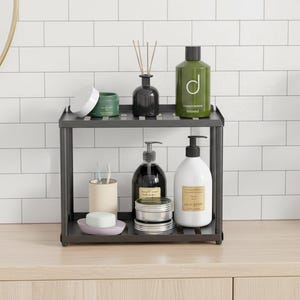 Może przedstawiać: Dwupoziomowa, ciemnoszara metalowa półka z różnymi niezbędnymi elementami łazienkowymi. Przedmioty obejmują zielone i czarne butelki, mały wazon z trzciną, uchwyt na szczoteczkę do zębów, mydło i małą roślinę. Półka stoi na jasnej drewnianej powierzchni.